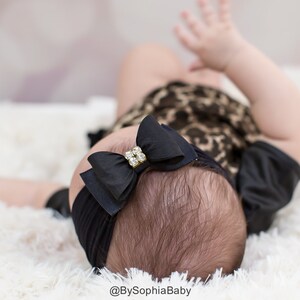 May include: A black baby headband with a large bow featuring a rhinestone center. The bow is centered on the baby's head, which is resting on a fluffy white surface. The baby is wearing a leopard print outfit.