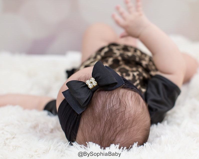 May include: A black baby headband with a large bow featuring a rhinestone center. The bow is centered on the baby's head, which is resting on a fluffy white surface. The baby is wearing a leopard print outfit.