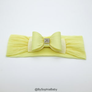 May include: A yellow headband with a large yellow satin bow and a rhinestone embellishment. The headband is made of stretchy fabric.
