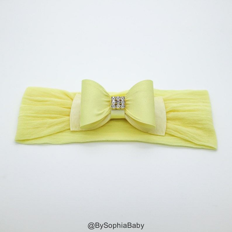 May include: A yellow headband with a large yellow satin bow and a rhinestone embellishment. The headband is made of stretchy fabric.