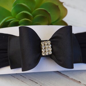 May include: A black baby headband with a large bow and a rhinestone center. The bow is made of a grosgrain ribbon and is attached to a soft, stretchy fabric band. The headband is a stylish accessory.