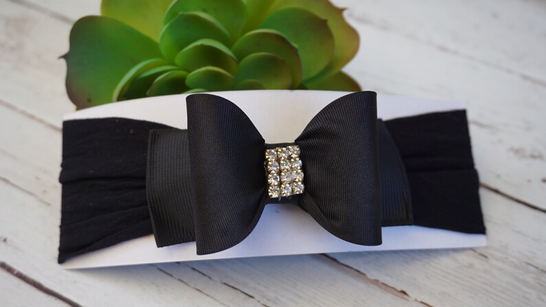 May include: A black baby headband with a large bow and a rhinestone center. The bow is made of a grosgrain ribbon and is attached to a soft, stretchy fabric band. The headband is a stylish accessory.