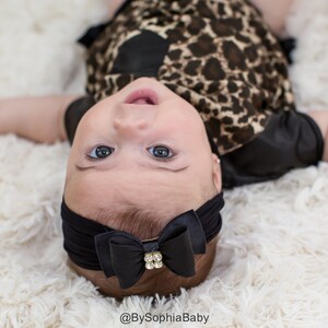 May include: A baby wearing a black headband with a bow and a rhinestone accent. The baby is wearing a leopard print outfit and is lying on a fluffy white surface. The baby is looking up at the camera.