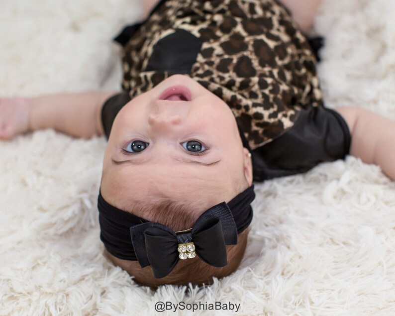 May include: A baby wearing a black headband with a bow and a rhinestone accent. The baby is wearing a leopard print outfit and is lying on a fluffy white surface. The baby is looking up at the camera.