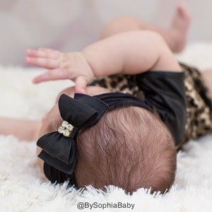 May include: A black baby headband with a layered bow and a rhinestone embellishment. The baby is wearing a black top and a leopard print outfit, lying on a white fluffy surface. The text "@BySophiaBaby" is visible.