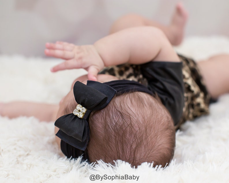 May include: A black baby headband with a layered bow and a rhinestone embellishment. The baby is wearing a black top and a leopard print outfit, lying on a white fluffy surface. The text "@BySophiaBaby" is visible.