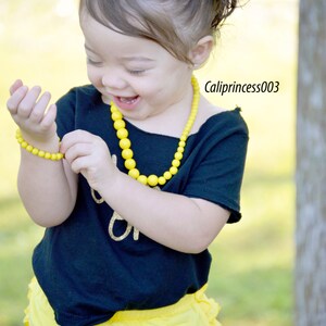 May include: A young girl wearing a black shirt with gold glitter lettering, yellow ruffled shorts, a yellow beaded necklace, and a yellow bow in her hair. She is smiling and holding a yellow beaded bracelet.