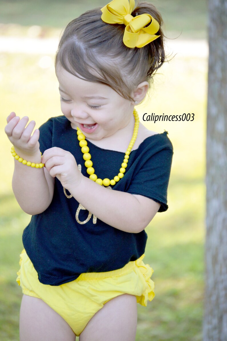 May include: A young girl wearing a black shirt with gold glitter lettering, yellow ruffled shorts, a yellow beaded necklace, and a yellow bow in her hair. She is smiling and holding a yellow beaded bracelet.