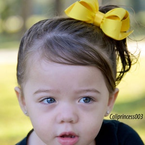 May include: A young girl with brown hair wearing a black shirt and a large yellow bow in her hair.