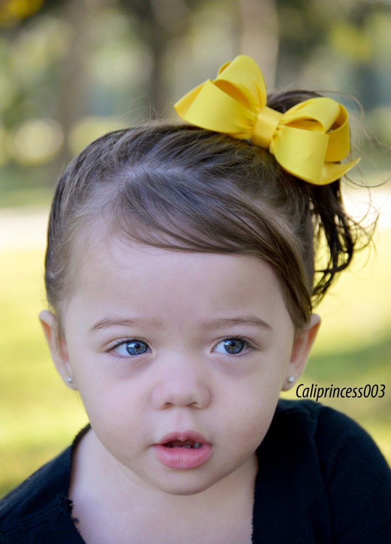 May include: A young girl with brown hair wearing a black shirt and a large yellow bow in her hair.