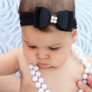 May include: A black bow headband with a gold and crystal embellishment is worn by a baby. The baby is also wearing a white pearl necklace. The text "@BySophiaHeadbands" is visible at the bottom of the image.