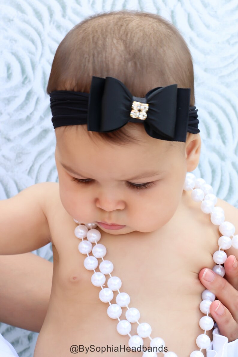 May include: A black bow headband with a gold and crystal embellishment is worn by a baby. The baby is also wearing a white pearl necklace. The text "@BySophiaHeadbands" is visible at the bottom of the image.