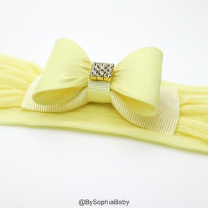 May include: A yellow headband with a large yellow satin bow and a gold rhinestone embellishment. The headband is made of stretchy fabric.