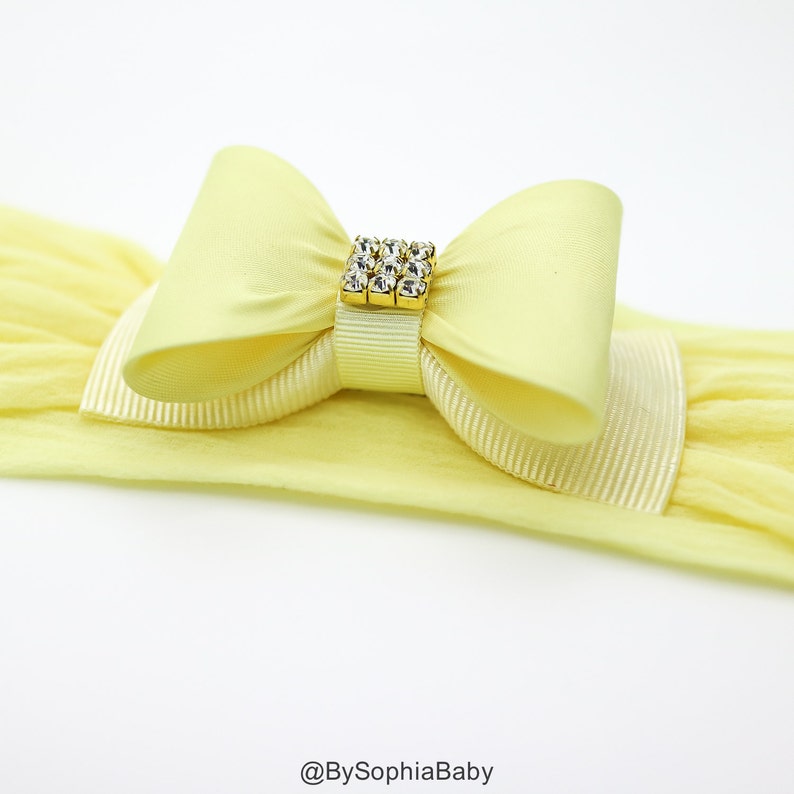 May include: A yellow headband with a large yellow satin bow and a gold rhinestone embellishment. The headband is made of stretchy fabric.