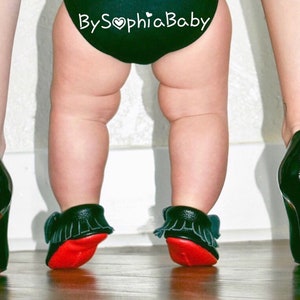 May include: A baby wearing black and red moccasins with fringe, standing in front of a person wearing black high heels with red soles. The baby's nappy has the text "By Sophia Baby" on it.