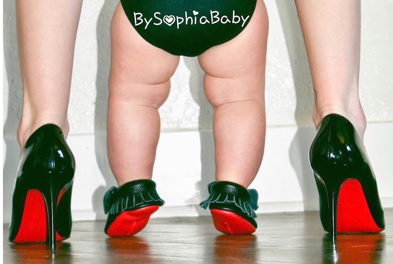 May include: A baby wearing black and red moccasins with fringe, standing in front of a person wearing black high heels with red soles. The baby's nappy has the text "By Sophia Baby" on it.