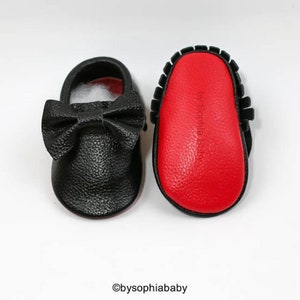 May include: A pair of black leather baby moccasins with a red sole. The moccasins have a bow on the front and the words "by Sophia Baby" are printed on the sole.