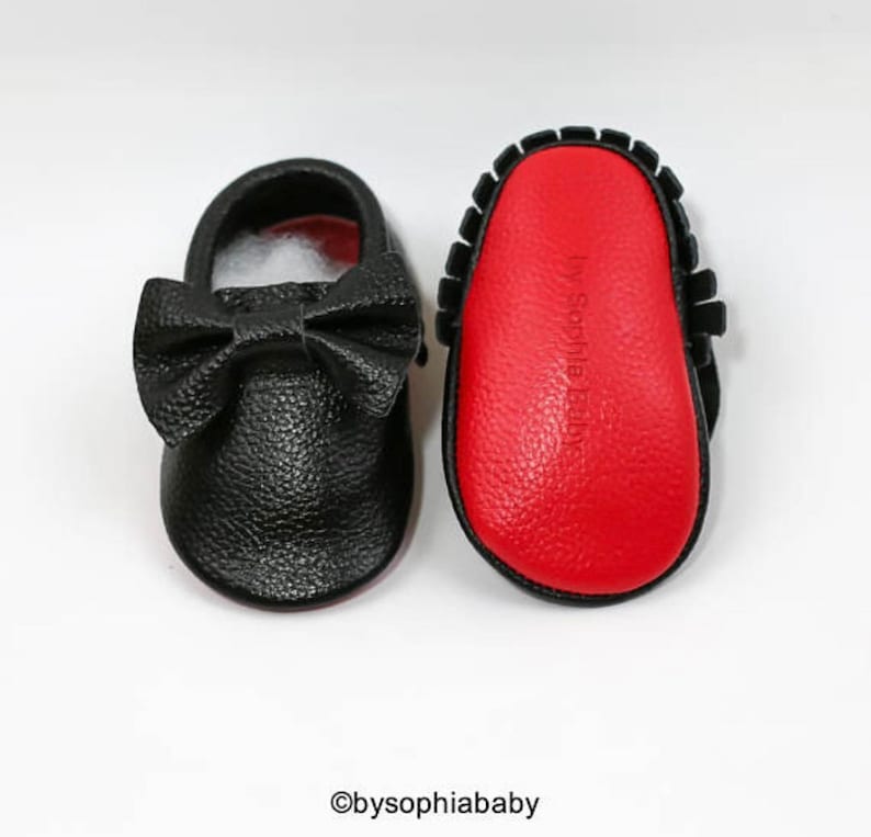 May include: A pair of black leather baby moccasins with a red sole. The moccasins have a bow on the front and the words "by Sophia Baby" are printed on the sole.