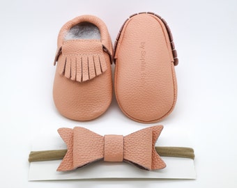 May include: A set of peach-coloured leather baby moccasins with fringe and a matching peach-coloured leather bow headband. The moccasins have the text "by Sophia Baby" on the back. The headband is on a brown elastic band.