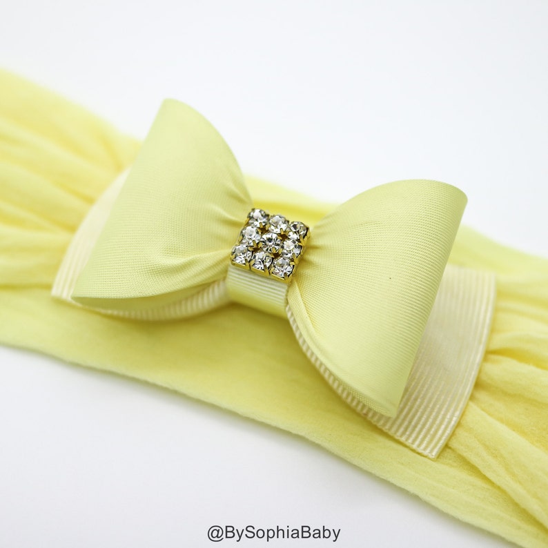 May include: A yellow headband with a large yellow satin bow and a gold and crystal embellishment. The headband is made of stretchy fabric.