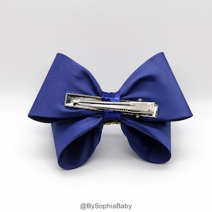 Op de afbeelding: Een grote blauwe satijnen strik met een zilveren alligatorclip. De strik is in een klassieke stijl geknoopt met twee lussen en een knoop in het midden.
