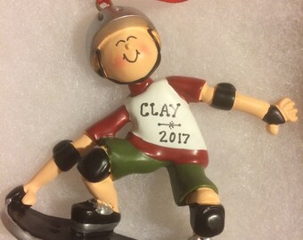 May include: A personalized Christmas ornament featuring a boy wearing a helmet and knee pads, riding a skateboard. The ornament is decorated with the name "CLAY" and the year "2017".