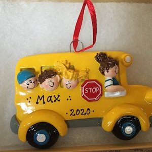 May include: A yellow school bus ornament with smiling children's faces peeking out of the windows. The bus has black wheels, a red stop sign, and the name "Max" and the year "2020" written on it. A red ribbon is attached for hanging.