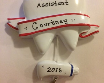 May include: A white dental assistant ornament shaped like a tooth, with the text "Dental Assistant" and "Courtney" on a red-trimmed banner. A small tube of toothpaste with "2016" and a red toothbrush hang below.