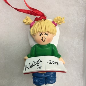 May include: A decorative ornament of a young girl holding a book. The girl has blonde hair in pigtails and is wearing a green shirt and blue jeans. The book reads "Adalyn - 2018". A red ribbon is attached for hanging.