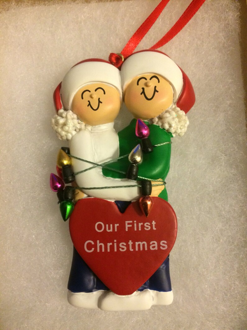 May include: A Christmas ornament featuring a couple holding a red heart with the text "Our First Christmas". The couple is dressed in winter clothing and is surrounded by colorful Christmas lights.