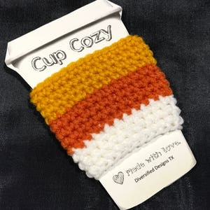 Op de afbeelding: Een witte bekerwarmer met een gehaakt ontwerp in oranje, wit en geel. De bekerwarmer heeft de tekst "Cup Cozy" aan de zijkant en de tekst "Made with Love, Diversified Designs TX" aan de onderkant.