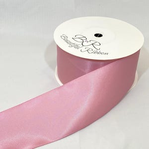 Puede incluir: Un carrete de cinta de raso rosa empolvado con el texto "B&R Beautiful Ribbon" impreso en la bobina de cartón blanco. La cinta está desenrollada, mostrando su textura suave y brillante.