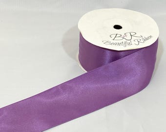 May include: A roll of lavender satin ribbon with the text "B R Beautiful Ribbon" printed on the white cardboard spool. The ribbon is unwound, showing its smooth, shiny texture. Perfect for crafts and gift wrapping.