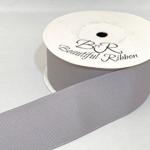May include: A spool of light gray grosgrain ribbon with a textured, ribbed surface. The white cardboard spool features the black printed text B R Beautiful Ribbon with a small pink bow icon between the letters B and R on the top edge.