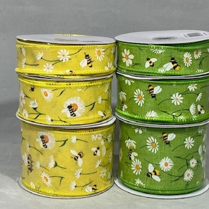 Op de afbeelding: Twee spoelen lint met een madeliefje- en bijenpatroon. Eén spoel is geel met witte madeliefjes en zwarte en gele bijen. De andere spoel is groen met witte madeliefjes en zwarte en gele bijen.