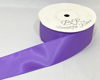 May include: A spool of purple satin ribbon with the text "B&R Beautiful Ribbon" printed on the white cardboard spool. The ribbon is a solid, vibrant purple color and is unrolled from the spool.