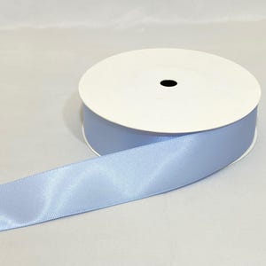 Puede incluir: Un rollo de cinta de raso azul claro. La cinta est&aacute; desenrollada, revelando su textura brillante. El rollo tiene un n&uacute;cleo de cart&oacute;n blanco con un agujero central negro. La cinta es de un azul pastel suave.