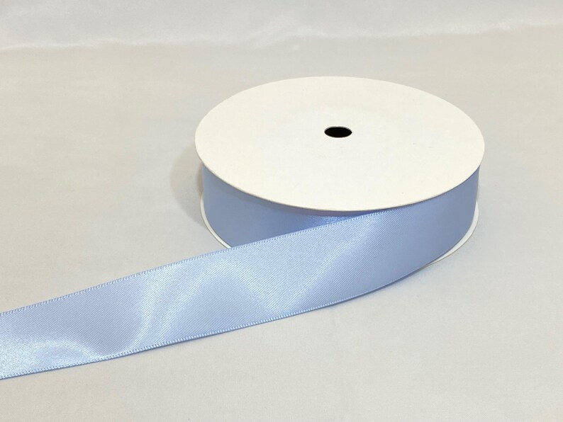 Puede incluir: Un rollo de cinta de raso azul claro. La cinta est&aacute; desenrollada, revelando su textura brillante. El rollo tiene un n&uacute;cleo de cart&oacute;n blanco con un agujero central negro. La cinta es de un azul pastel suave.