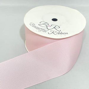 Op de afbeelding: Een spoel lichtroze grosgrain lint met een textuur van ribbels. Het witte kartonnen middenstuk is voorzien van de zwart gedrukte tekst B&R Beautiful Ribbon met een klein roze strikicoontje tussen de letters B en R op het label.