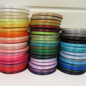 May include: Three vertical stacks of satin ribbon rolls in a wide variety of colours, including white, yellow, orange, pink, red, green, blue, purple, and black. Each roll is tightly wound, displaying a smooth, reflective texture and sheen.