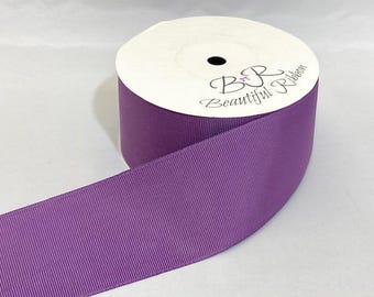 May include: A spool of solid purple grosgrain ribbon with a ribbed texture. The white cardboard center features the black printed text BR Beautiful Ribbon with a small pink bow icon. The ribbon is wound tightly around the central spool.
