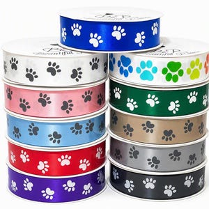 May include: A collection of ten spools of satin ribbon stacked in two columns. Each ribbon features a repeating pattern of paw prints. The colours include white, pink, light blue, red, purple, royal blue, rainbow, green, tan, grey, and black.
