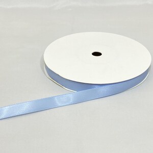 Puede incluir: Un rollo de cinta de raso azul claro. La cinta est&aacute; desenrollada, revelando su textura suave y su color. El rollo es blanco con un borde azul, y la cinta es de un azul pastel suave.