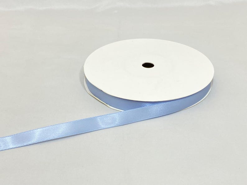 Puede incluir: Un rollo de cinta de raso azul claro. La cinta est&aacute; desenrollada, revelando su textura suave y su color. El rollo es blanco con un borde azul, y la cinta es de un azul pastel suave.