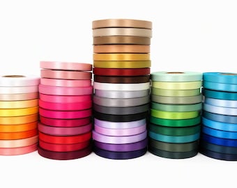 May include: Four vertical stacks of satin ribbon rolls in a wide spectrum of colors, including shades of pink, orange, yellow, red, brown, white, gray, black, purple, green, and blue, arranged in a gradient pattern against a plain white background.