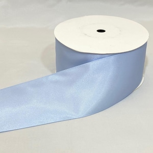 Puede incluir: Un rollo de cinta de raso azul claro. La cinta se desenrolla, revelando una textura suave y brillante. El rollo es blanco con un agujero central negro. Ideal para manualidades, envolver regalos y proyectos decorativos.