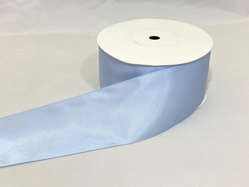 Puede incluir: Un rollo de cinta de raso azul claro. La cinta se desenrolla, revelando una textura suave y brillante. El rollo es blanco con un agujero central negro. Ideal para manualidades, envolver regalos y proyectos decorativos.