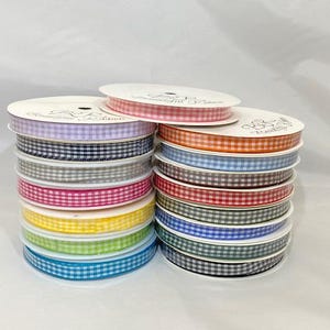 Könnte beinhalten: Stapel von Vichy-Bandspulen in verschiedenen Farben, darunter Rosa, Orange, Rot, Grün, Blau und Gelb. Das Band ist ordentlich auf weißen Spulen aufgewickelt, ideal für Bastelarbeiten und Geschenkverpackungen.