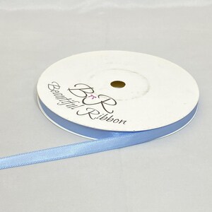 Puede incluir: Un carrete de cinta de raso azul claro. El carrete de cart&oacute;n blanco tiene el texto "Beautiful Ribbon" impreso. La cinta desenrollada est&aacute; plana sobre una superficie clara.
