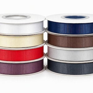 Könnte beinhalten: Zwei Stapel mit je vier Rollen geripptem Ripsband. Der linke Stapel enthält weiße, cremefarbene, rote und dunkelviolette Bänder. Der rechte Stapel enthält marineblaue, braune, hellgraue und anthrazitfarbene Bänder auf weißen Kunststoffspulen.